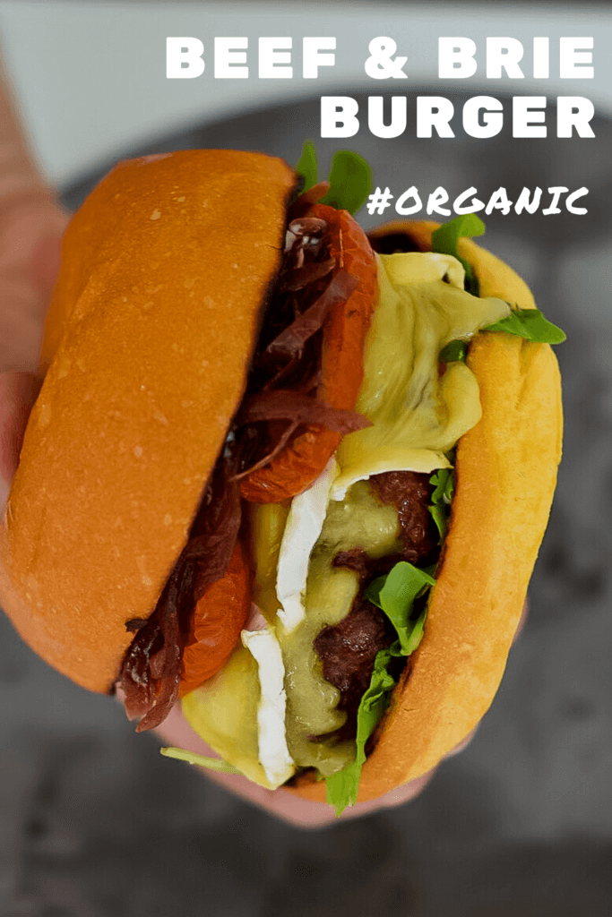 someone holding an organic beef burger with brie, roasted tomatoes & onion marmalade
