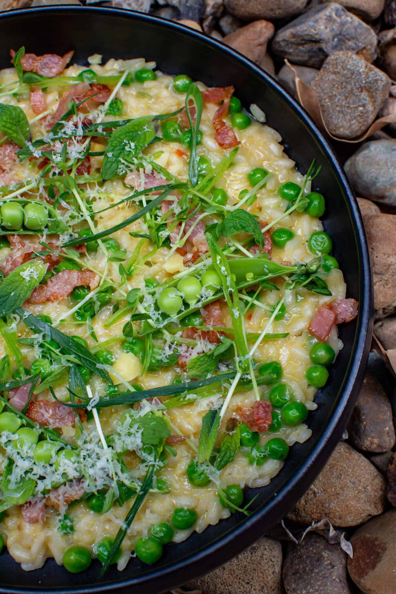 pea & bacon risotto in a black bowl on top of loads of stones
