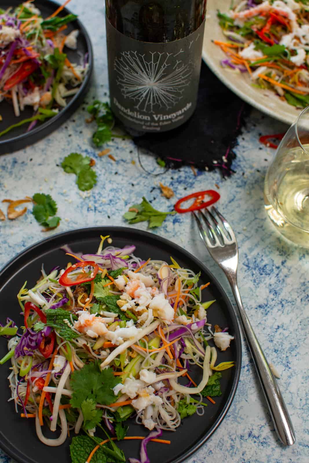 spicy crab salad on a black plate with a fork and a bottle of dandelion riesling on table