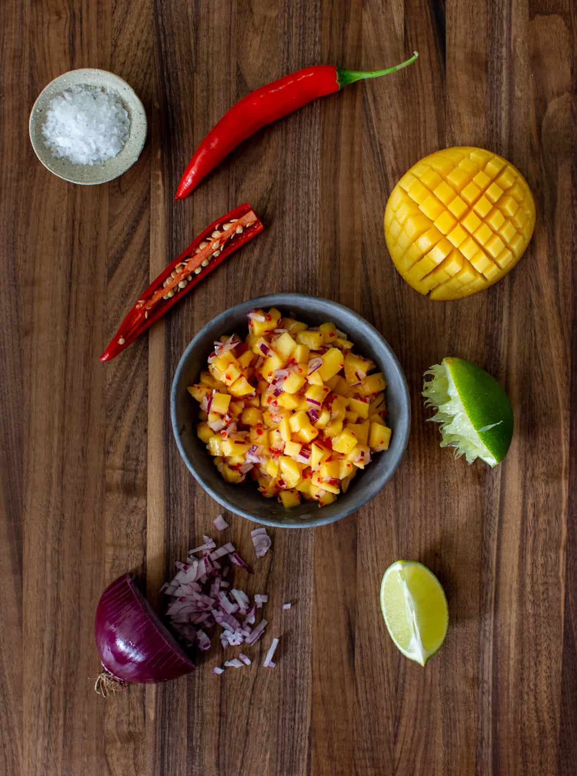 spicy mango salsa in a bowl on a wooden chopping board with salsa ingredients beside it