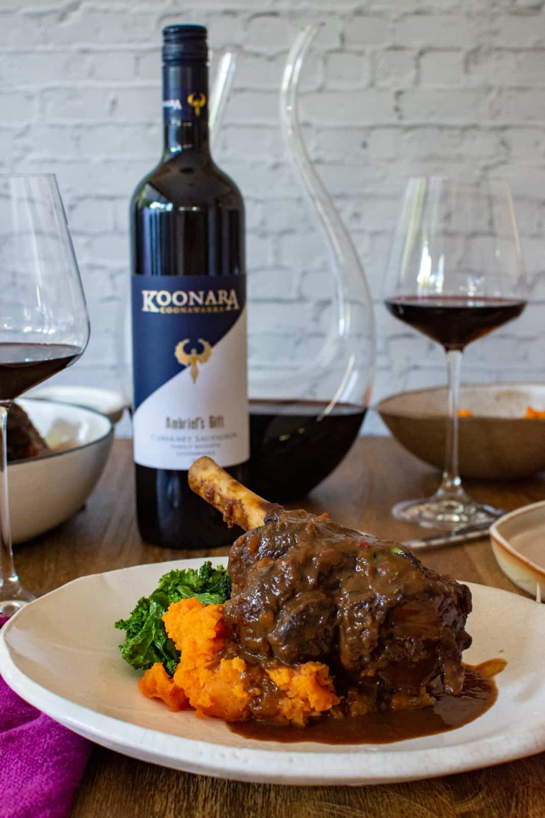 chocolate & chili braised lamb shanks on a white plate, koonara cabernet sauvignon bottle and glass of wine