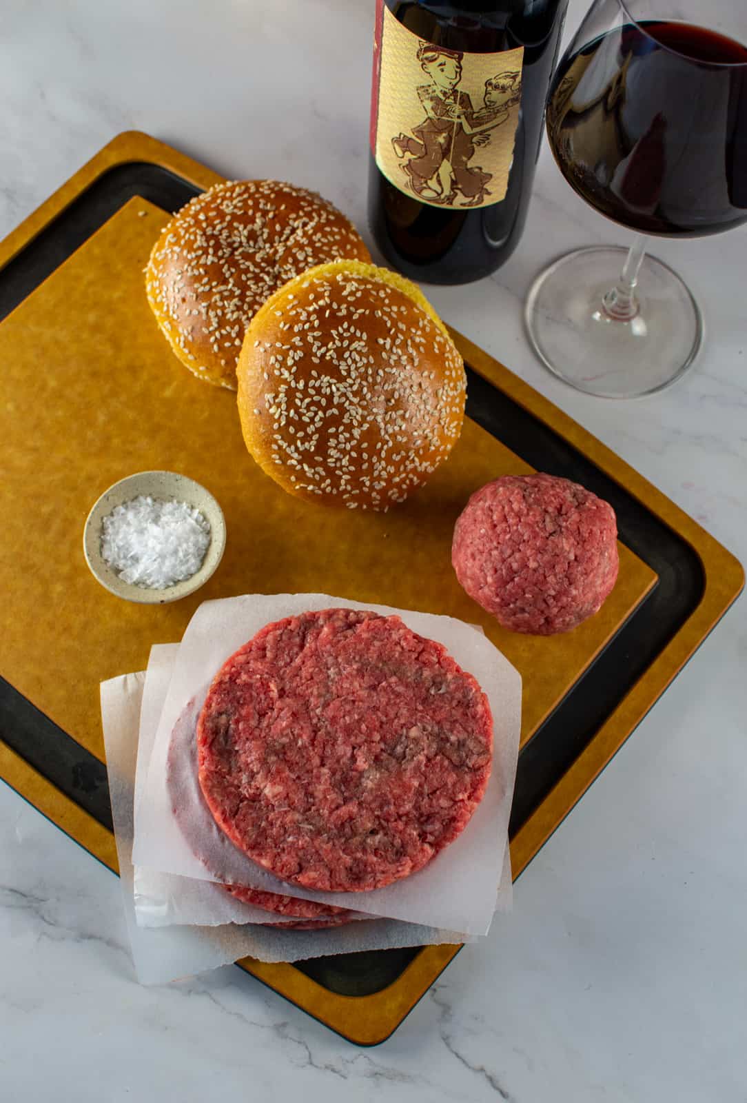 beef patties, salt, brioche buns on a chopping board with red wine beside it
