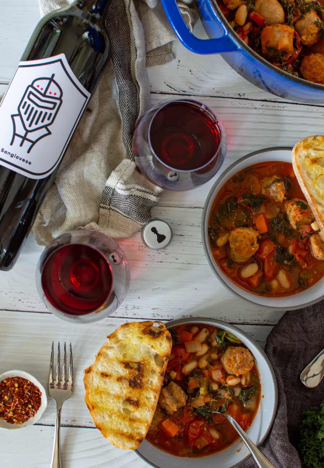 tuscan sausage stew in blue casserole dish, bowls of stew and pawn wine co red wine on a white table