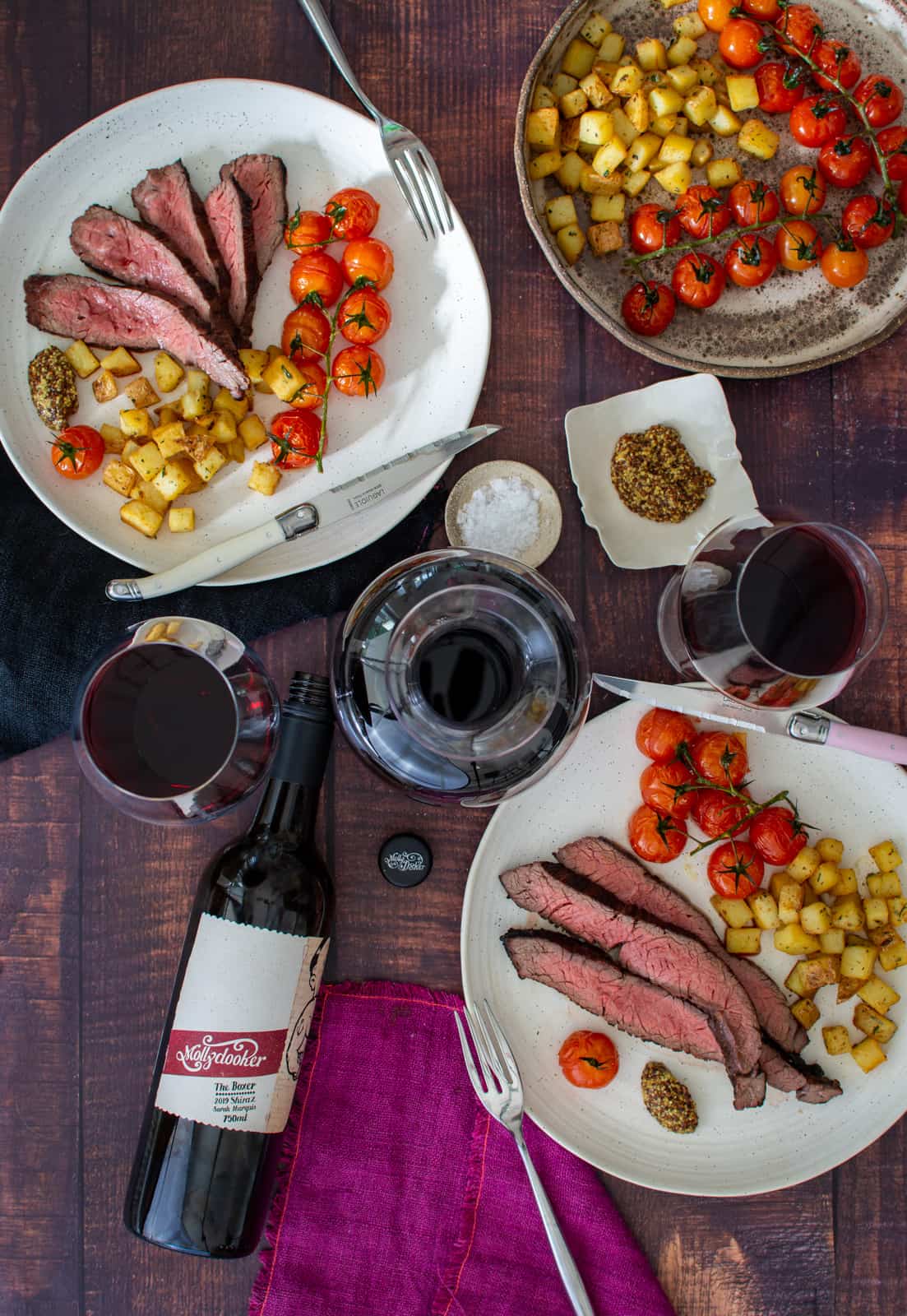 Birds eye view of bavette steak, potatoes & tomatoes on a plate with mollydooker red wine on table