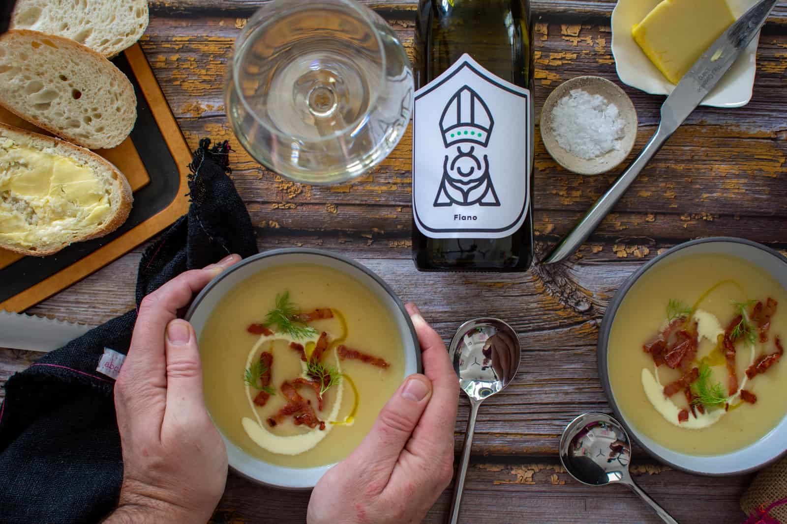 Two bowls of parsnip soup on a table with someone holding one of the bowls, bread, white wine, butter and salt also on the table 