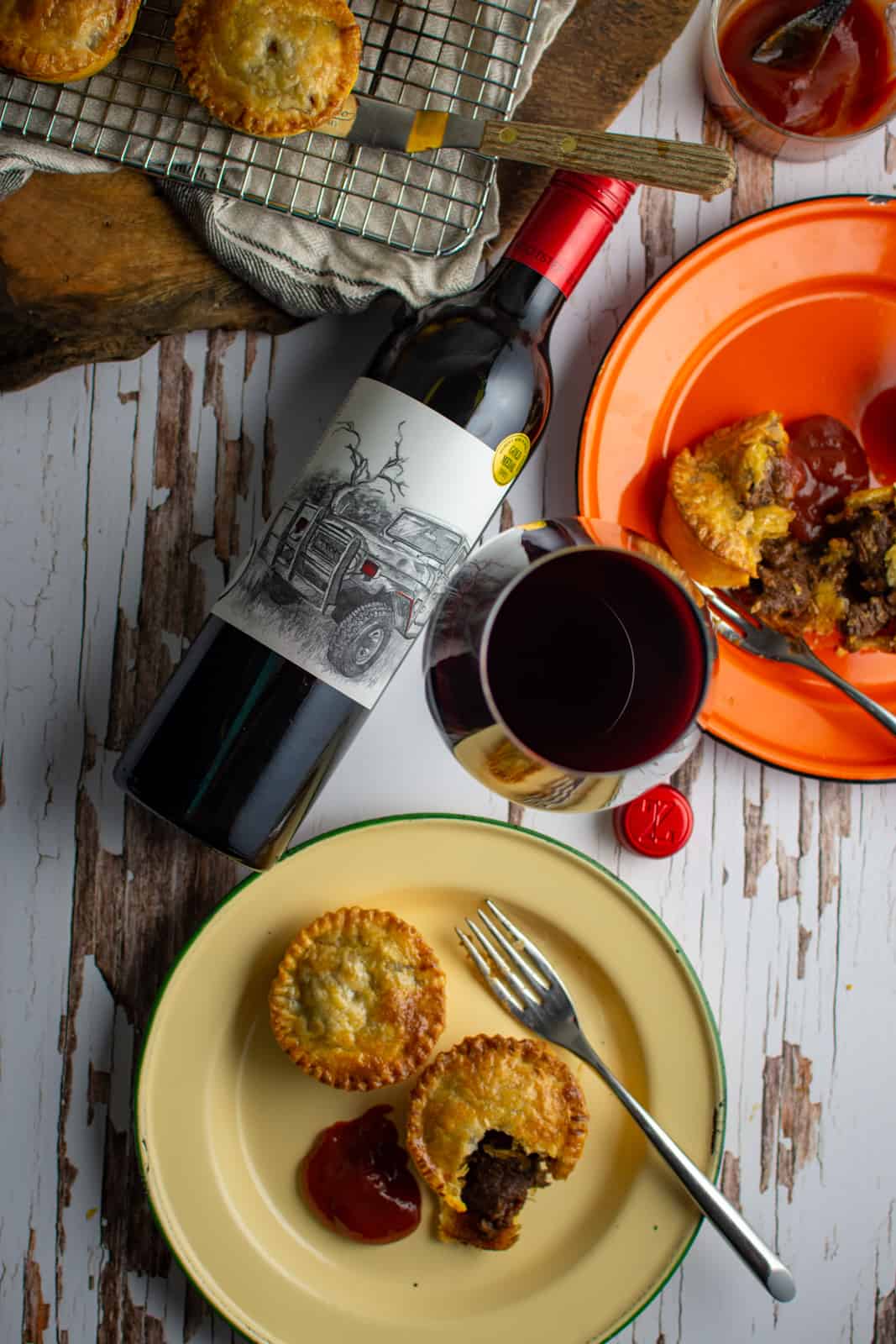 chunky beef pies on plates, red wine lying on its side and more pies on a rack