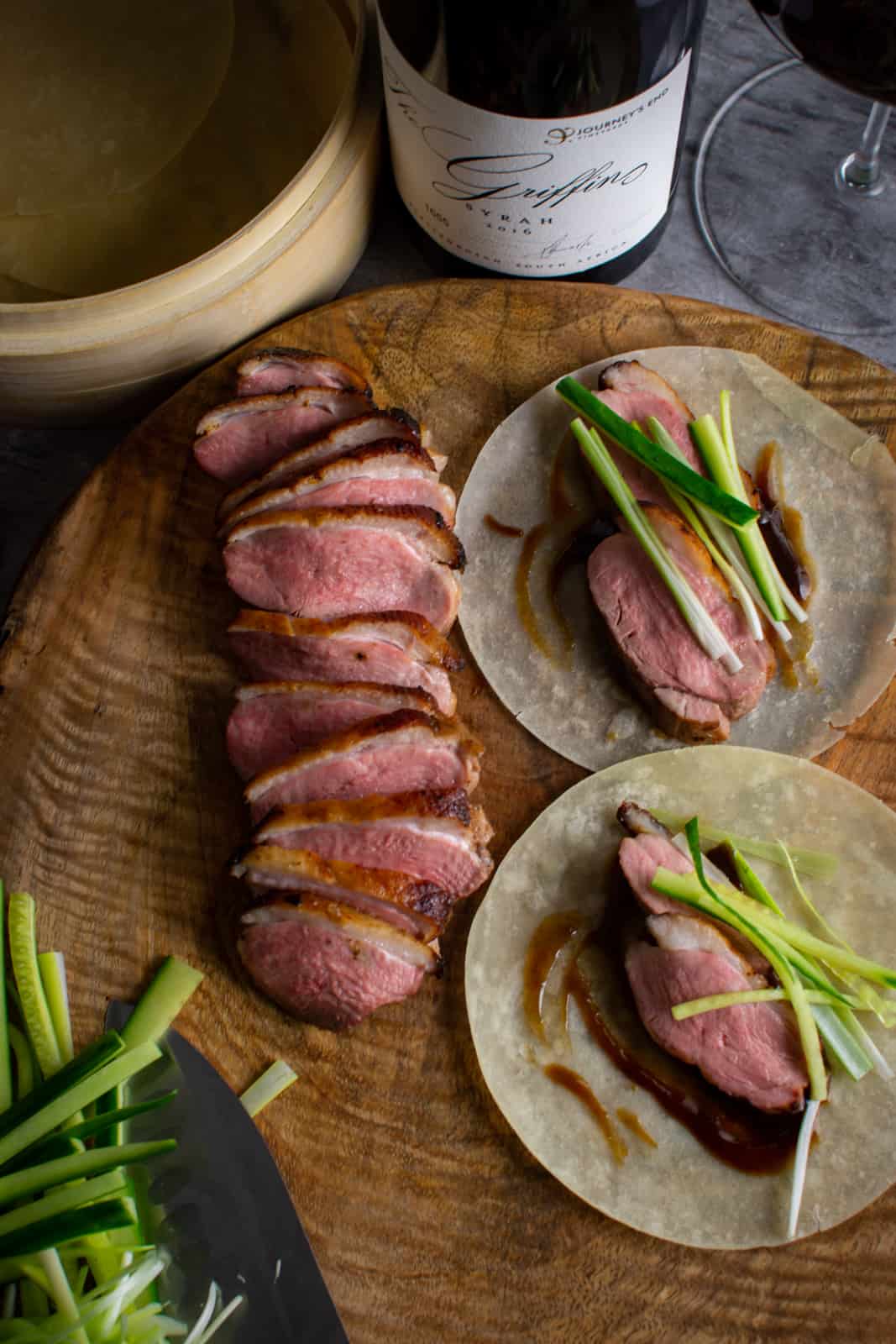 peking duck pancakes on wooden chopping board, cooked duck breast next to them with red wine too