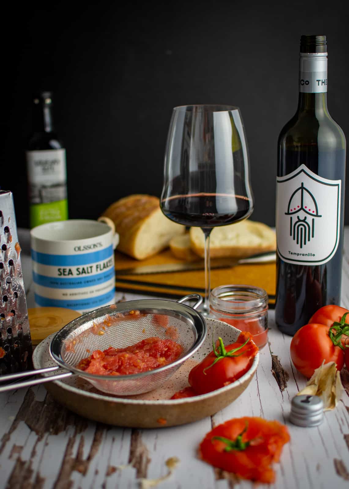 grated tomatoes in a bowl with a sieve. Red wine, wine glass, salt and bread in background