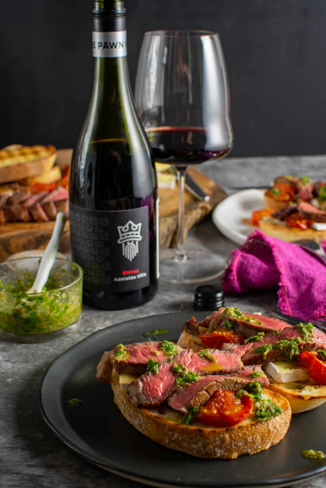 steak crostini on a black plate, plate, salsa and steak in background with a bottle of wine too