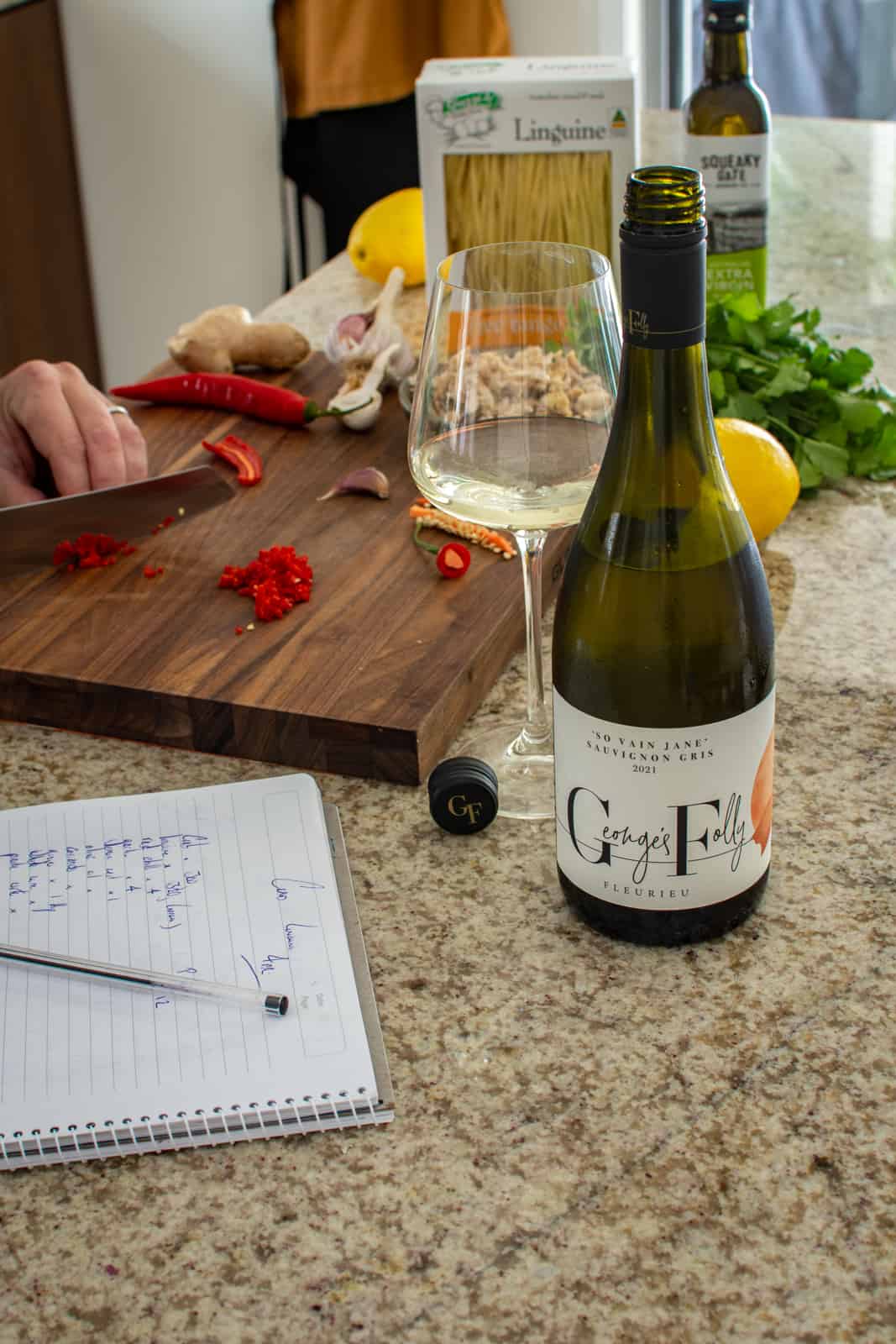 someone chopping chilli in a kitchen with food on counter and a bottle of wine too