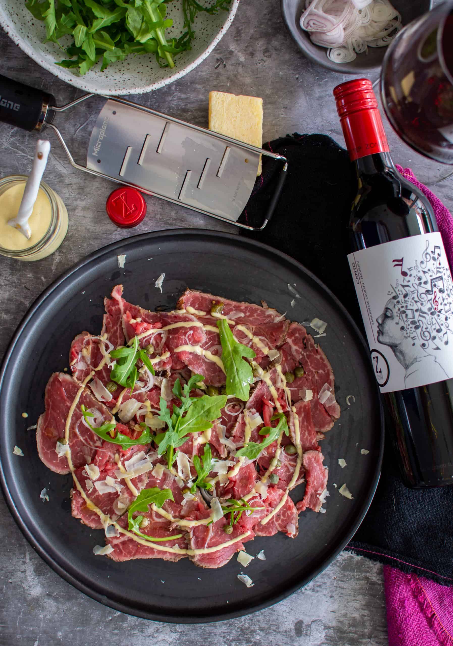 beef carpaccio on a black plate, parmesan cheese, rocket and a bottle of red wine