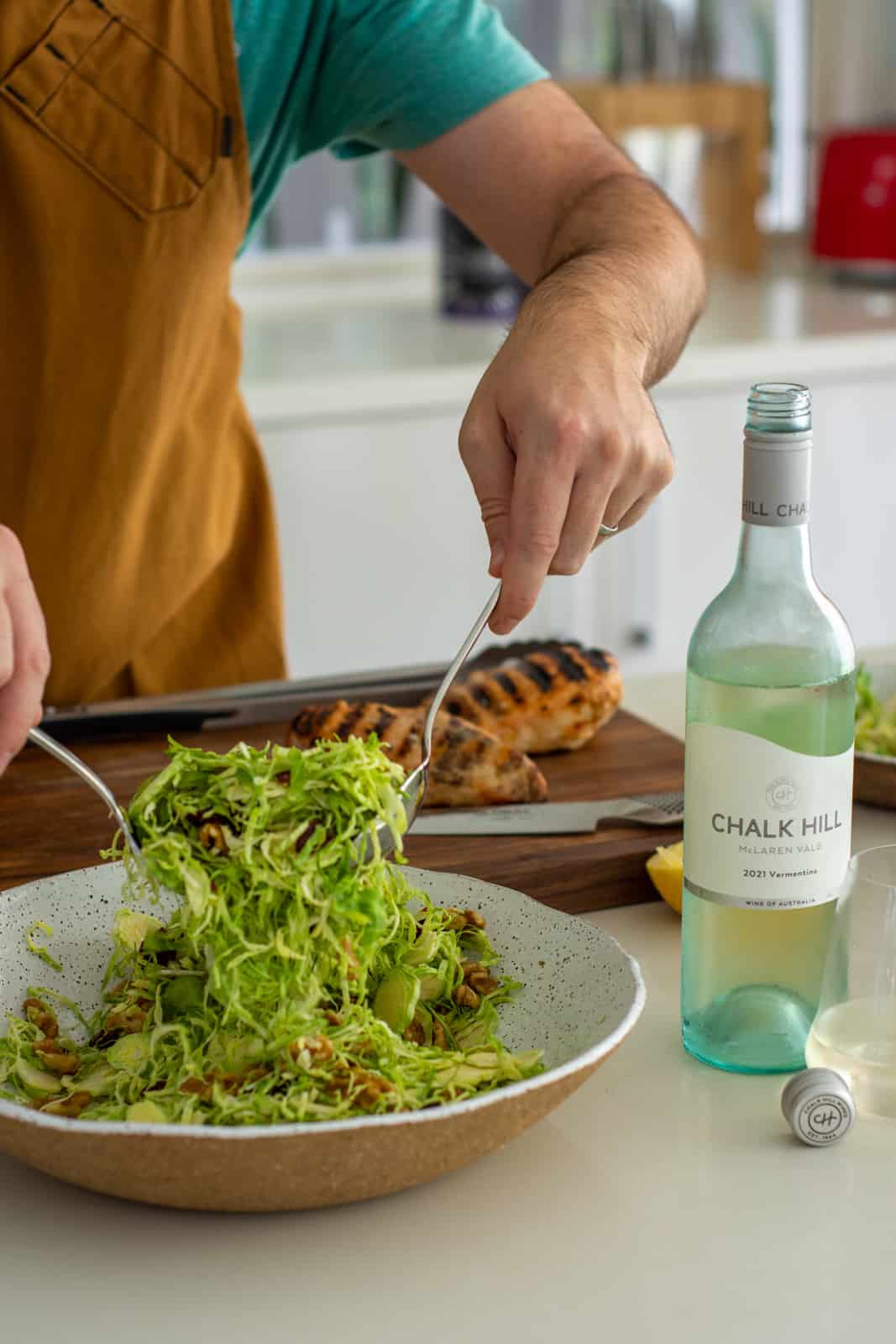 Someone tossing shaved brussels sprout salad in the kitchen, grilled chicken on chopping board behind