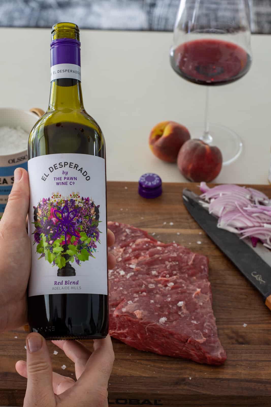 someone holding a bottle of red wine with steak on a chopping board beside