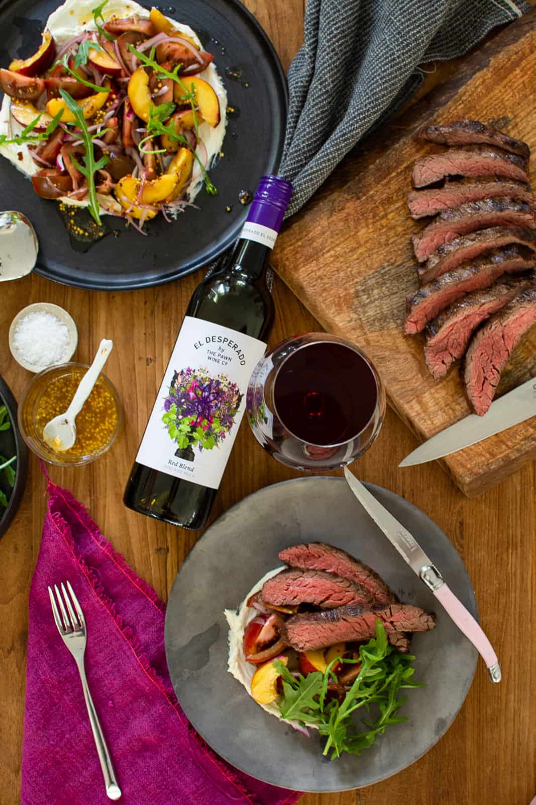 Birds-eye view of bavette steak and peach tomato salad, red wine on table and steak on chopping board