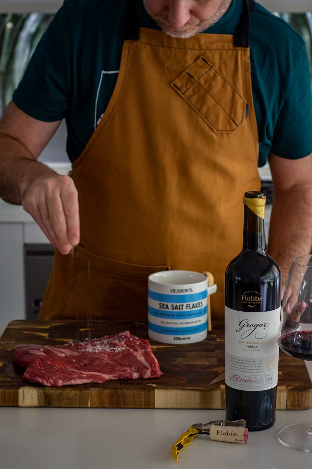 someone seasoning a steak and a bottle of red wine next to chopping board 