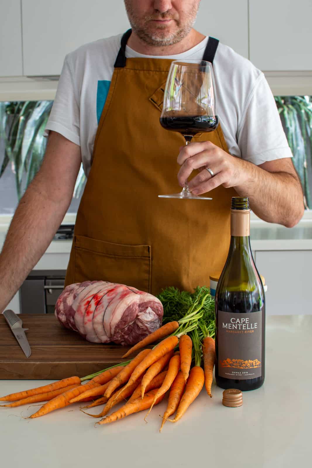 someone holding a glass of wine in the kitchen with boneless lamb leg and other ingredients on counter