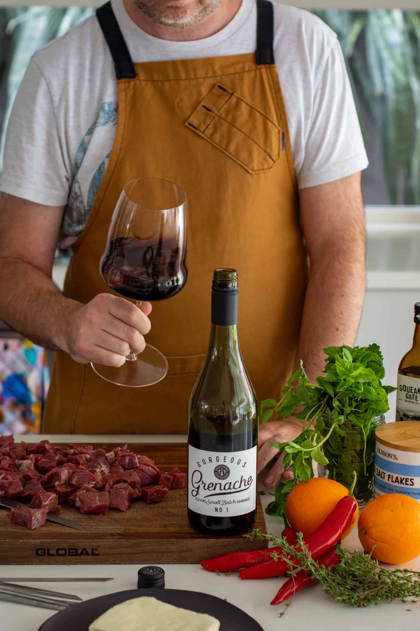 Someone swirling a glass of wine in the kitchen with food on chopping board and on kitchen counter top