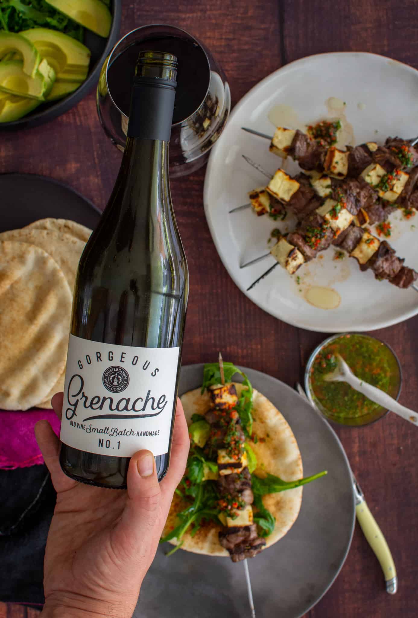 Someone pouring red wine with grilled lamb skewers below