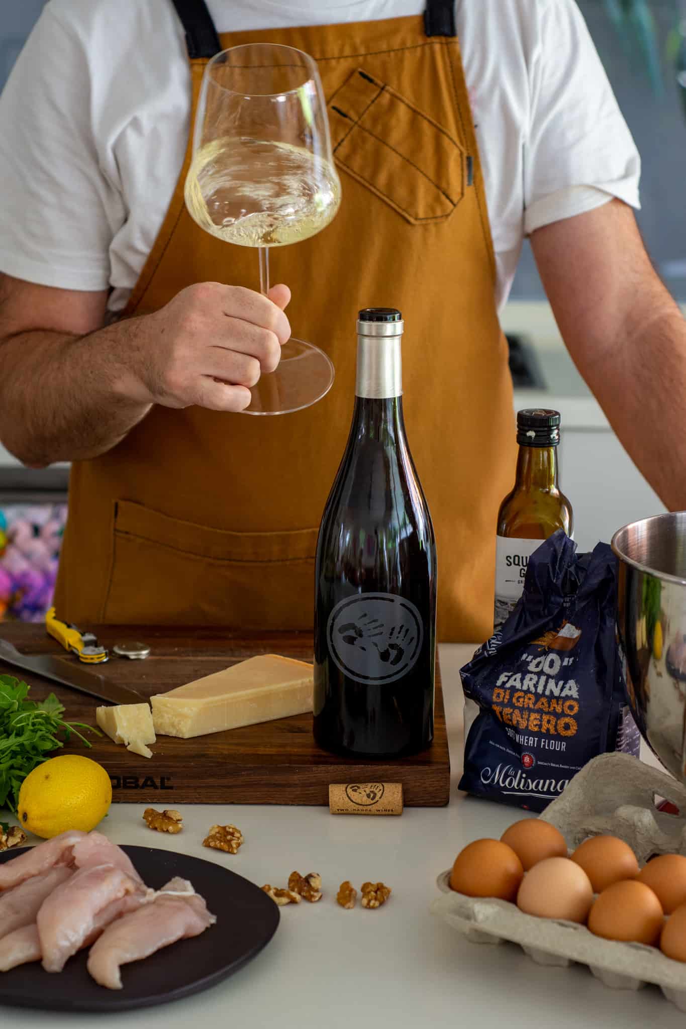 Someone swirling white wine in the kicthen with pasta al pesto ingredients on the counter