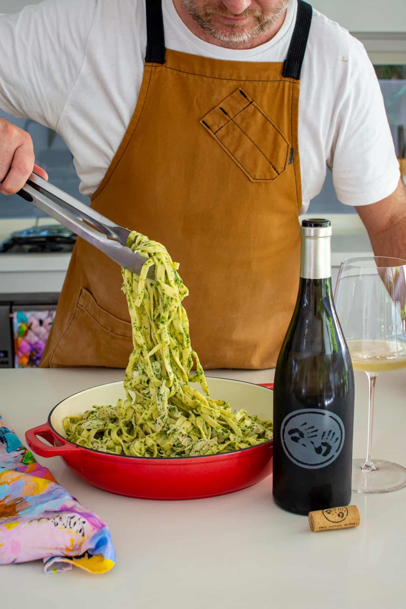 someone picking pasta from a red casserole dish using tongs. White wine beside them
