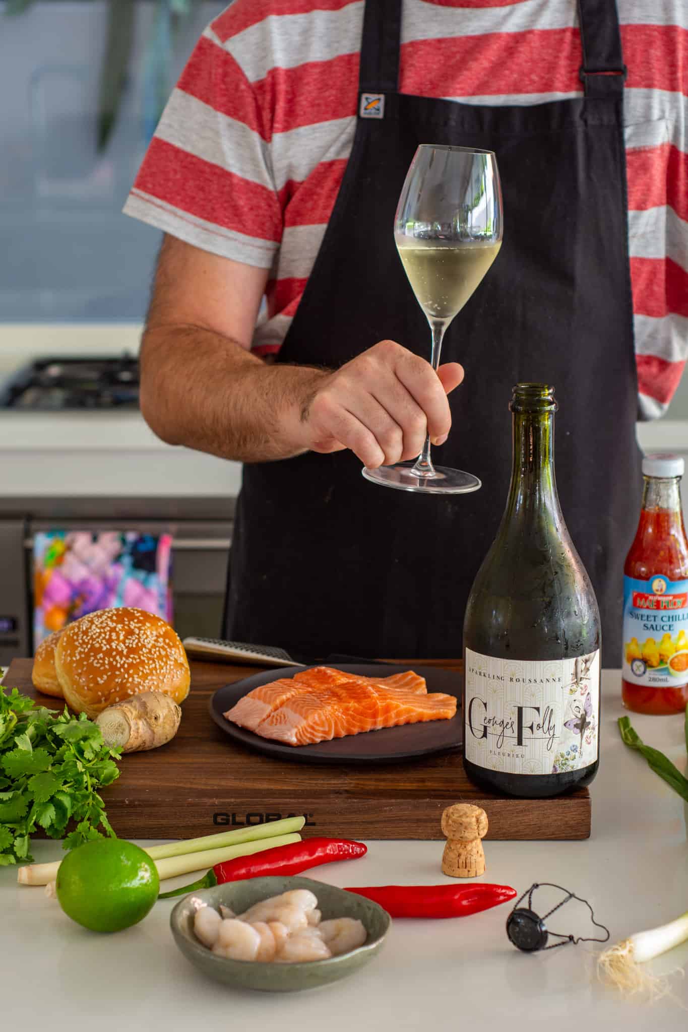 Someone holding a glass of sparkling wine with ingredients to make fish burger on kitchen counter top