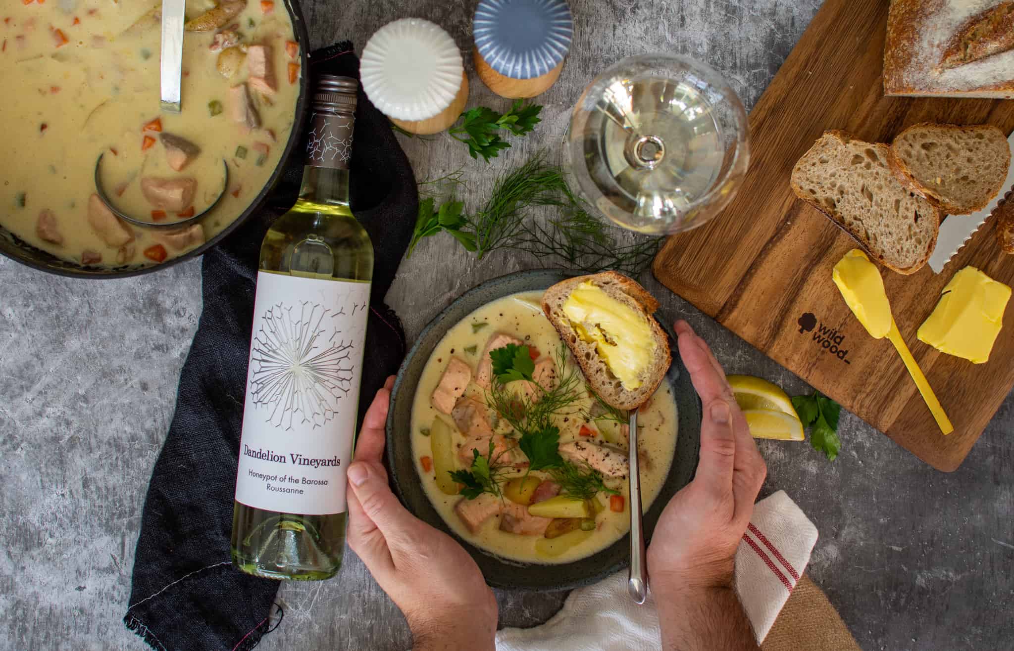 Birdseye view of someone holdng a bowl of chowder with bread, wine and saucepan of chowder next to them.