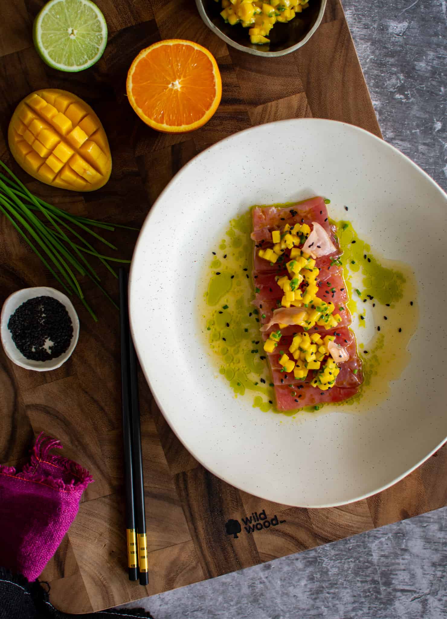 birds eye view of tuna crudo, mango chilli salsa on a white plate with citrus and mango on chopping board beside it