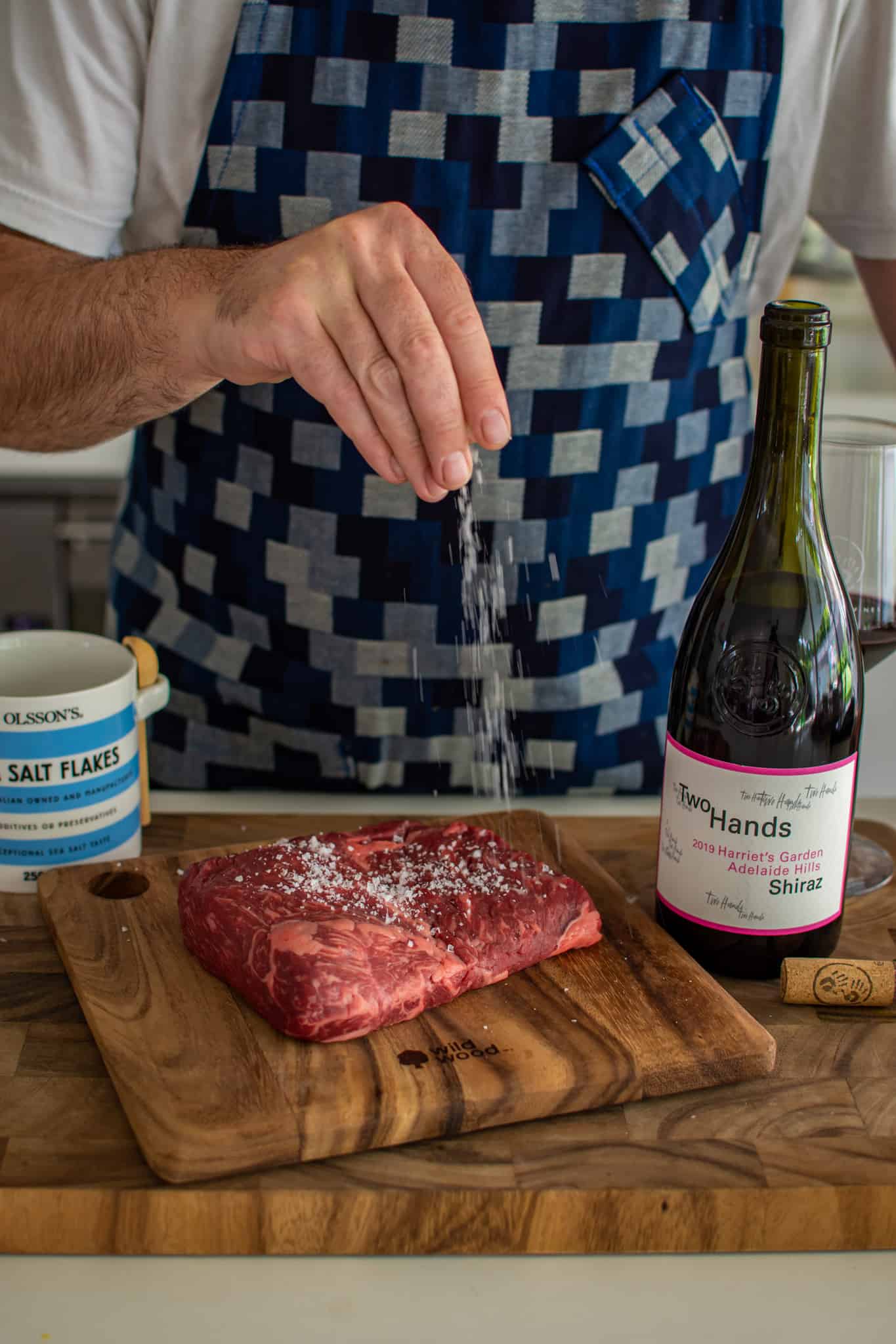 someone salting a steak in the kitchen with a bottle of wine next to the chopping board