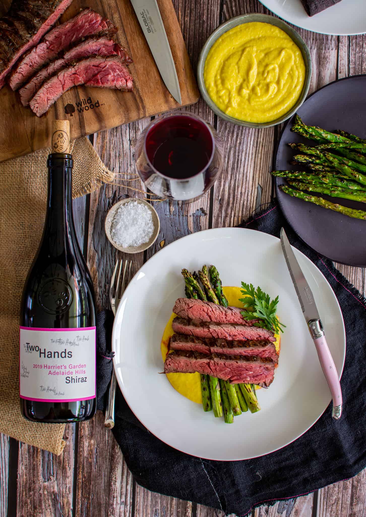 Birdseye view of grilled bavette steak, corn puree and asparagus on a plate. A bottle of Two Hands wine next to plate