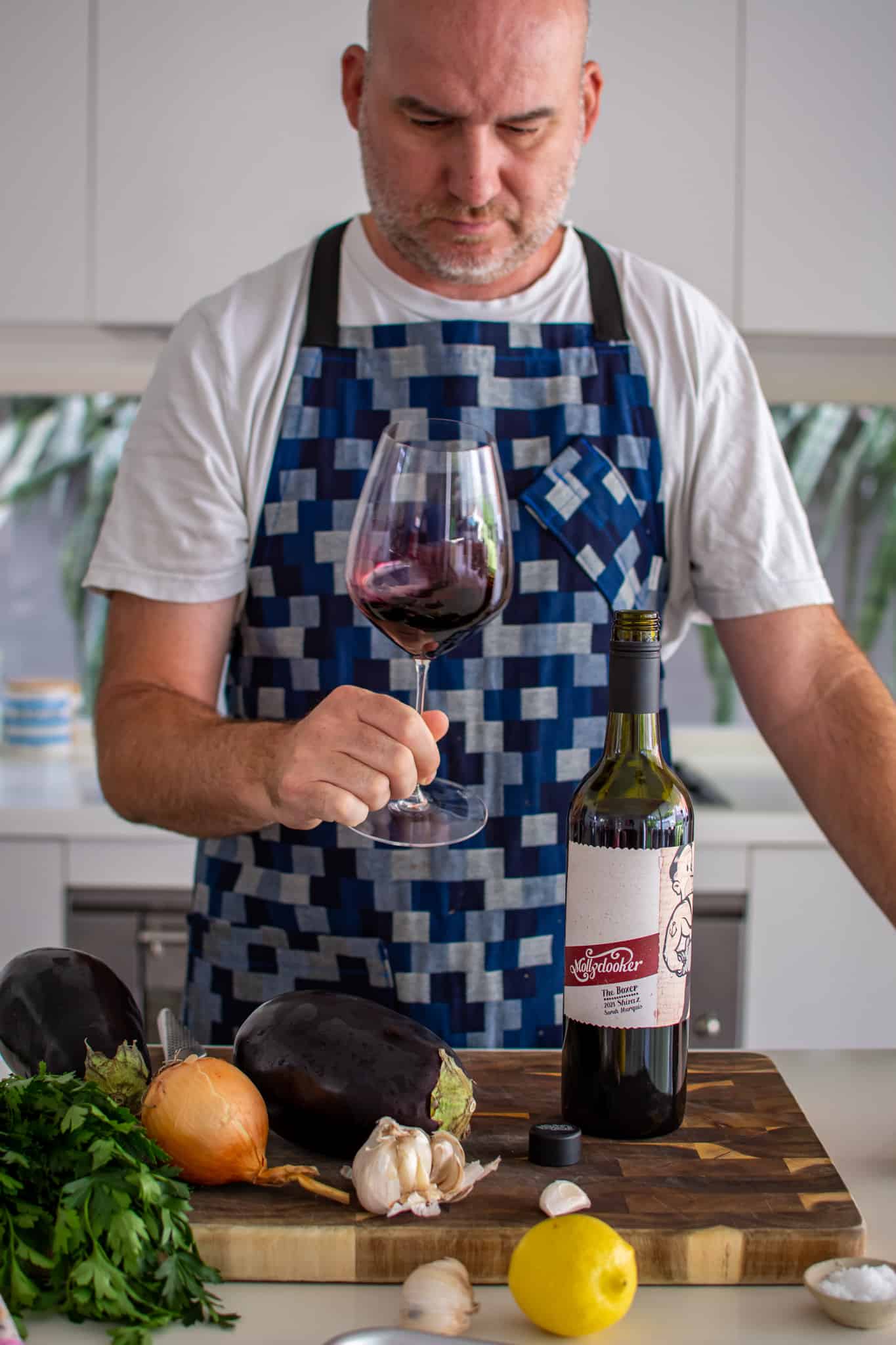 someone in the kitchen holding a glass of red wine with a bottle on the counter top and ingredients on chopping board