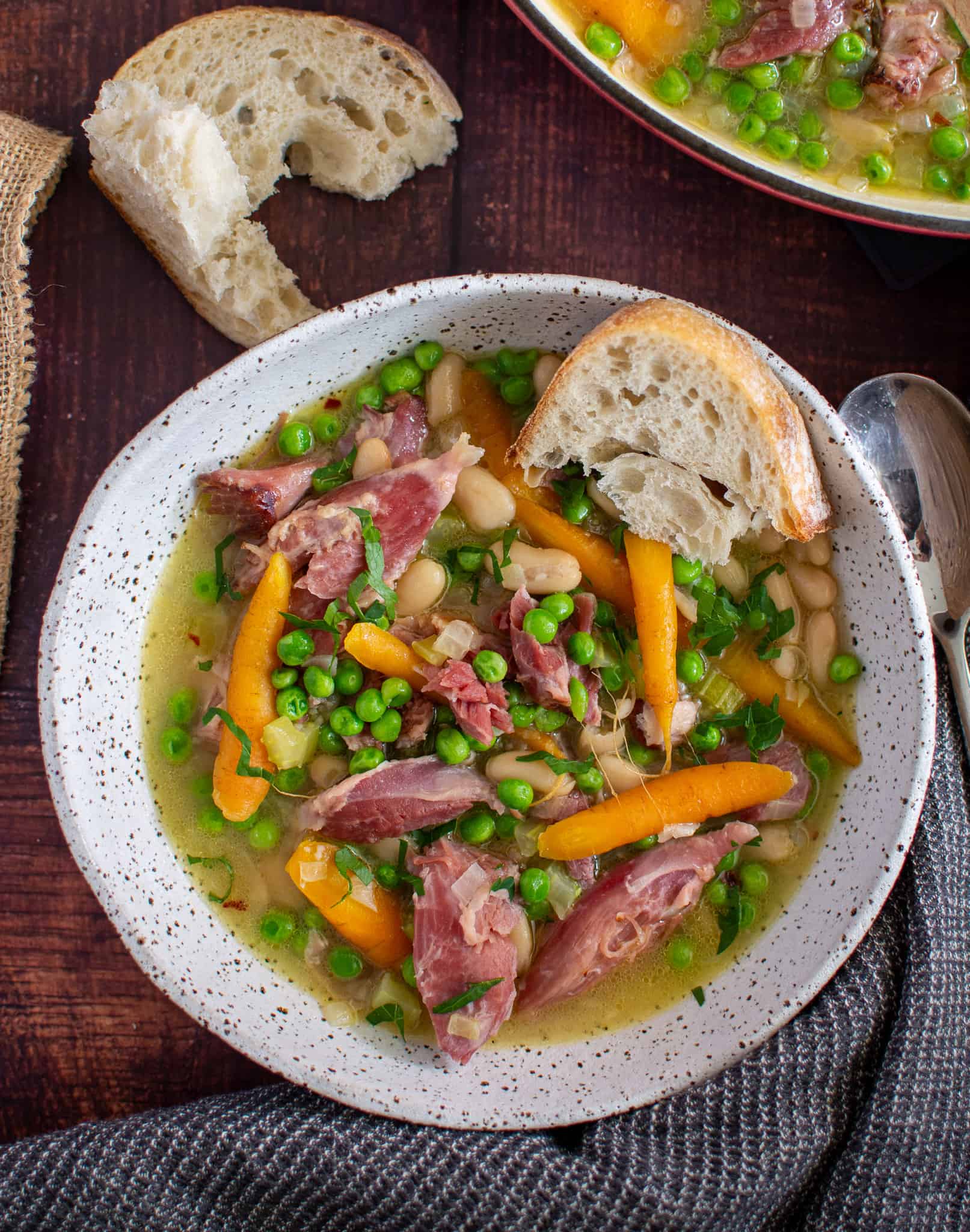 A bowl of ham hock stew with some bread lying on the table next to it