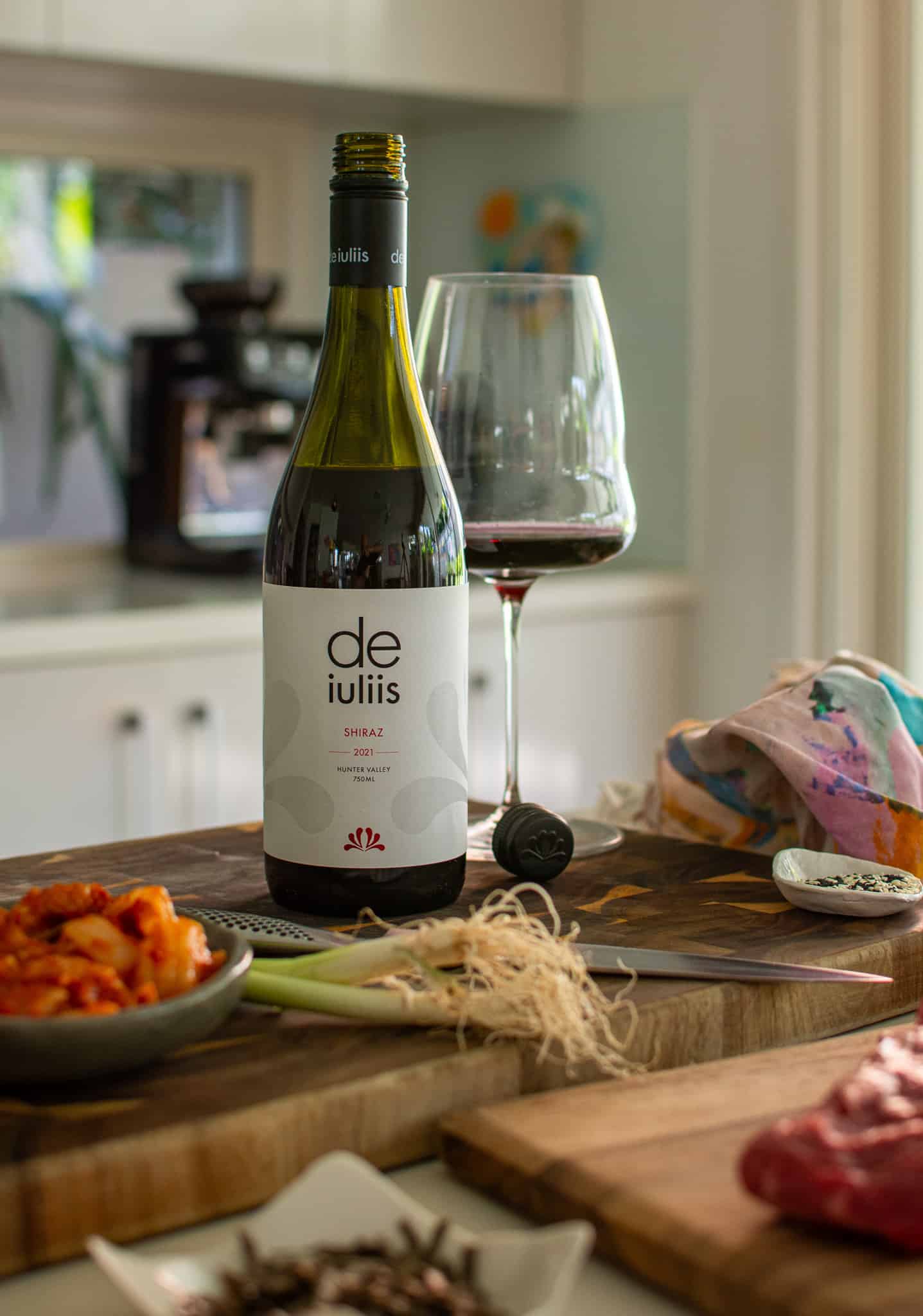 a bottle of De iuliis shiraz with wine glass on a chopping board with food lying beside it on the counter