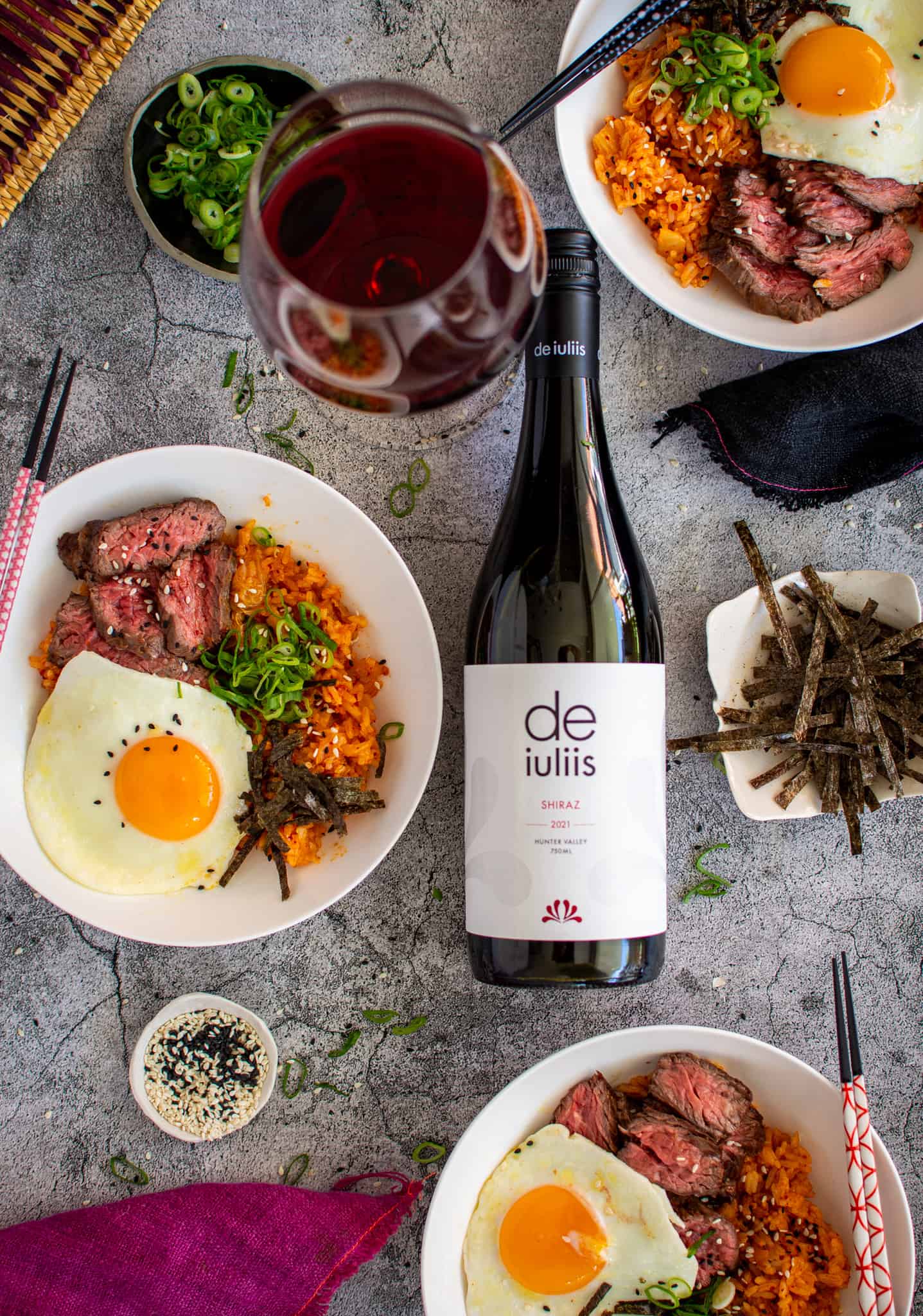 birdseye view of bowls of Korean fried rice, a bottle of De iuliis shiraz, wine glass and napkins