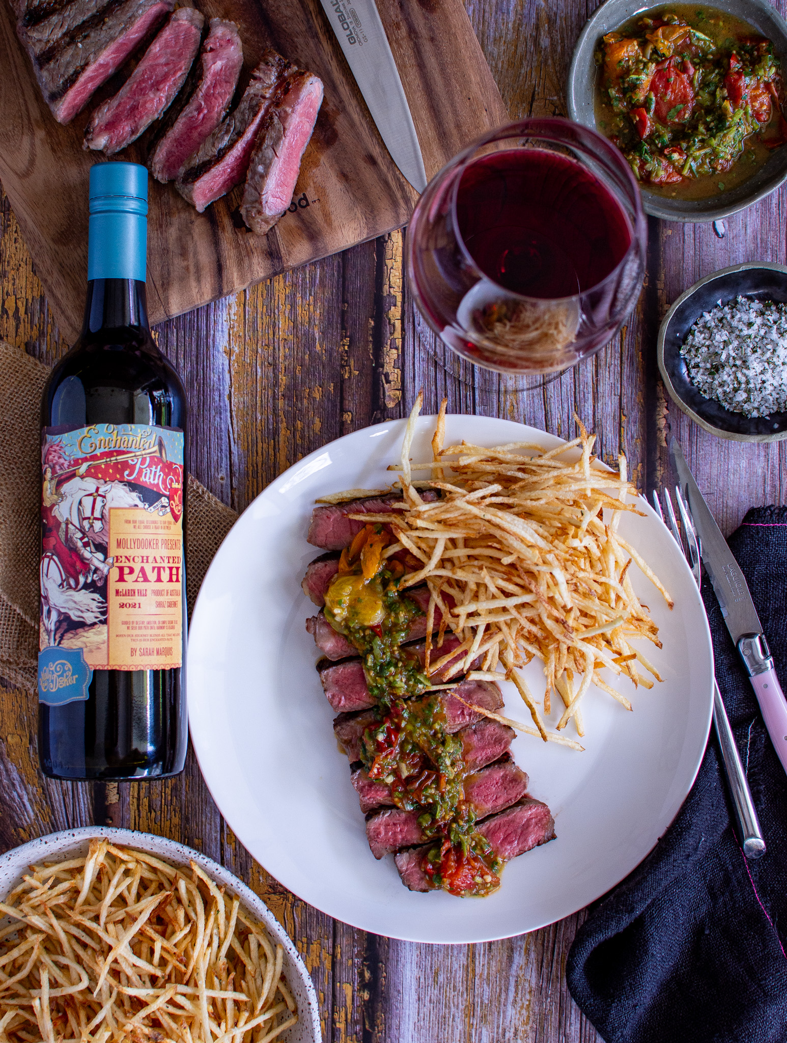 birdseye view of steak with tomato chimichurri on a plate, bottle of mollydooker wine, wine glass and sliced steak on chopping board