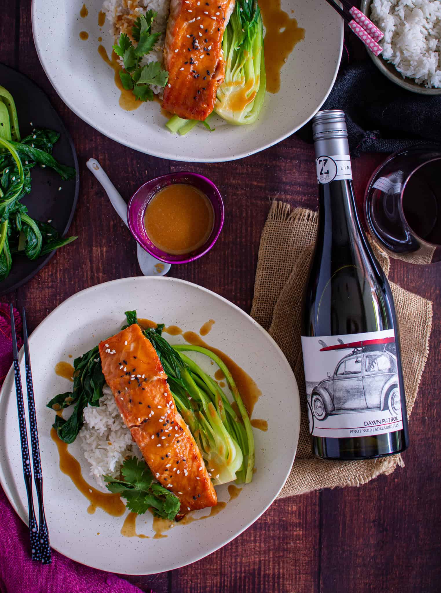 birds eye view of 2 plates of miso ginger salmon with rice and asian greens. Bottle of red wine lying next to the plates
